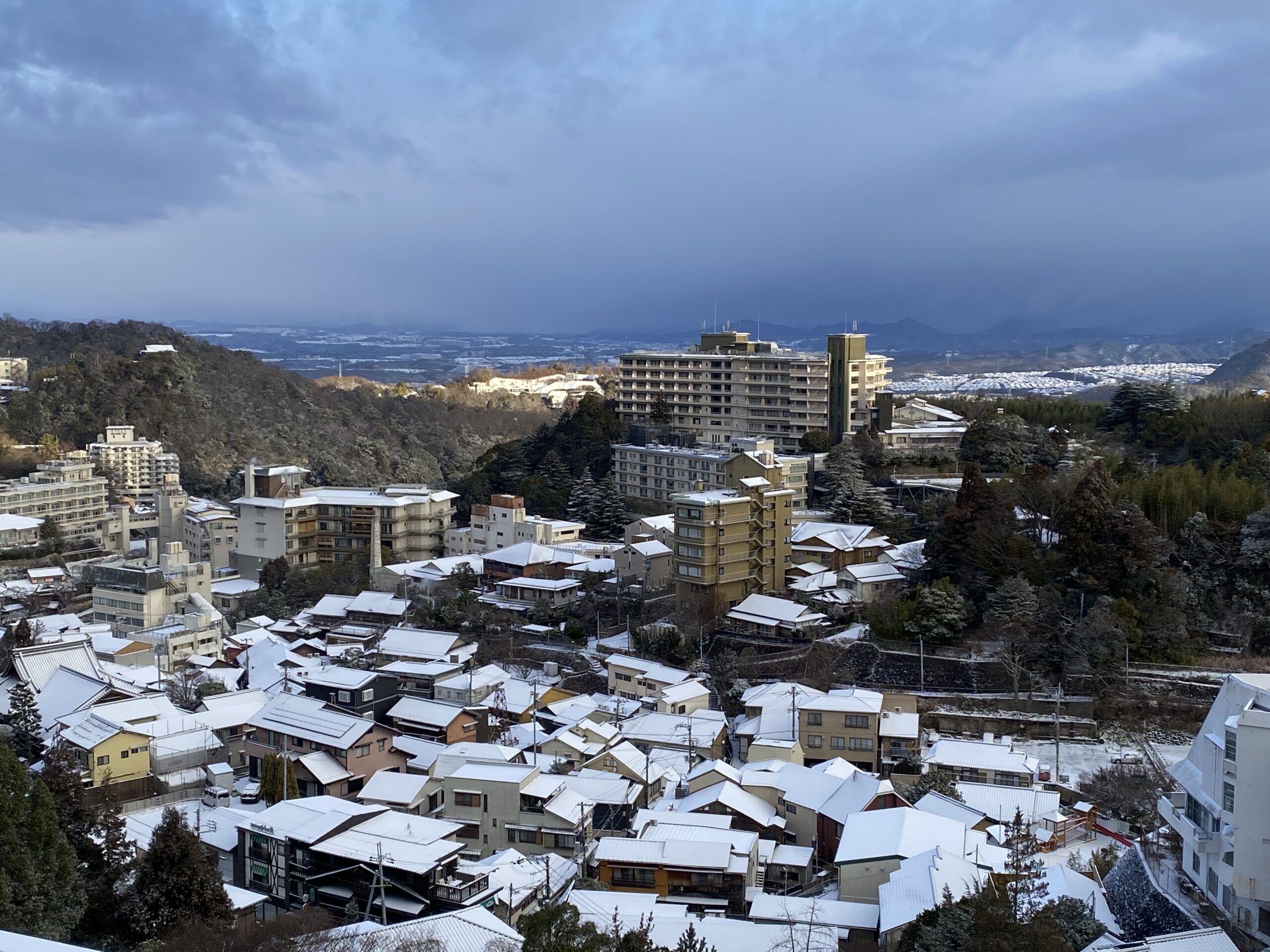 雪が一瞬止んだ朝の有馬温泉の町並み。白い屋根と山並みの上に光と雲が広がる冬の景色